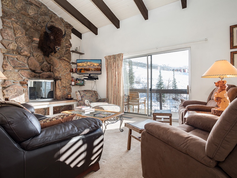Cozy up in your mountain retreat with soaring beamed ceilings, a striking stone fireplace, and plush seating overlooking snowy slopes—pure alpine comfort awaits.