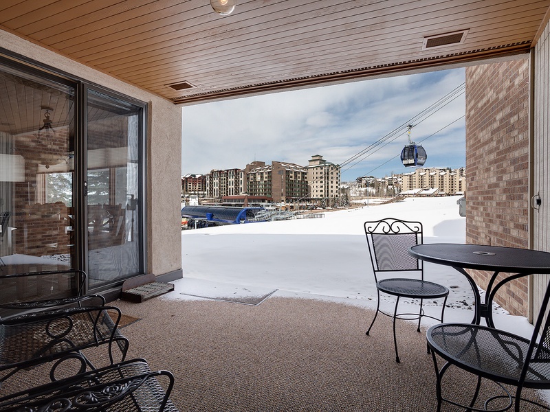 A covered patio with seating overlooks a snowy ski resort base area, offering views of slopes, gondola access, and surrounding village buildings amid winter scenery.