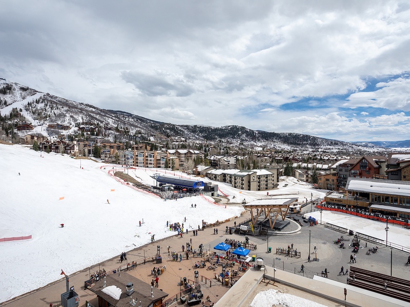 Aerial view of a mountain resort village nestled against snow-covered slopes, featuring ski facilities, lodges, and a bustling base area with shops and restaurants.