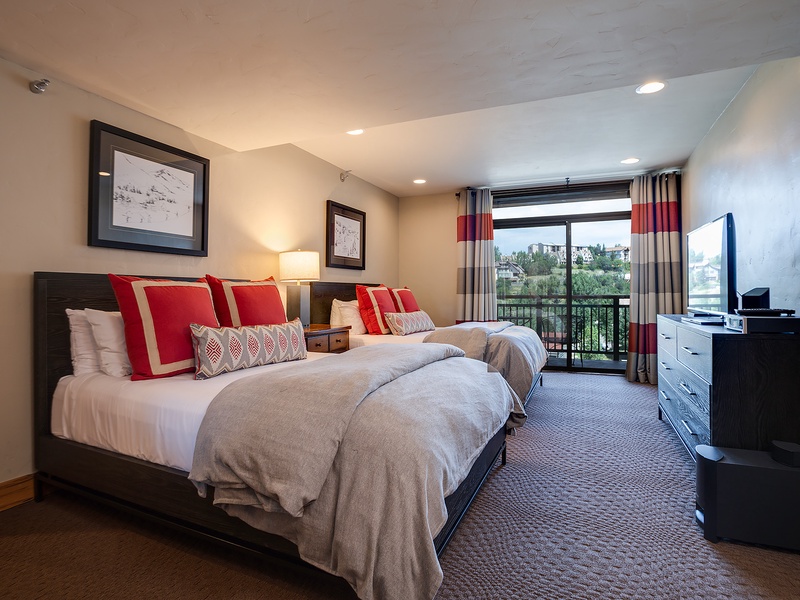 Two plush beds dressed in soft linens and bold red accents await you in this spacious bedroom, complete with a balcony overlooking the surrounding neighborhood and modern entertainment amenities.