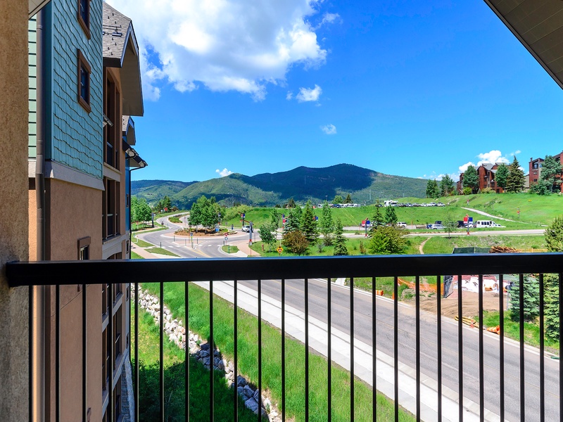 Panoramic mountain views stretch across rolling green hills and forests, framed by a balcony overlooking the surrounding resort area and distant peaks.