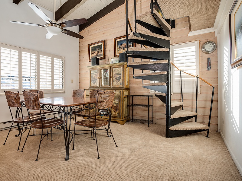 Picture yourself gathered around this inviting dining table, sunlight streaming through plantation shutters, with a charming spiral staircase leading to your cozy loft retreat above.