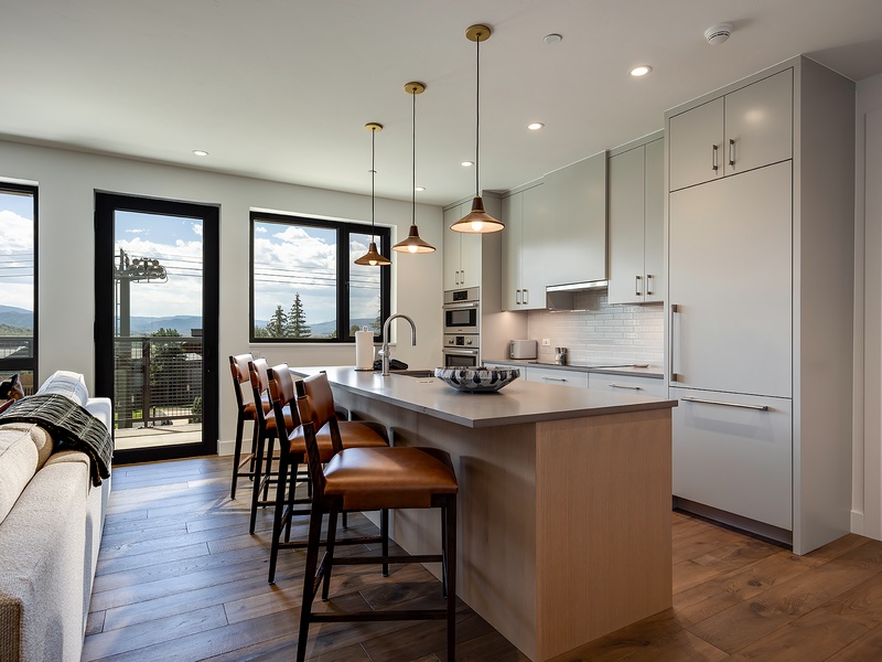 Step into your open-concept kitchen with sleek white cabinetry, a spacious island for gathering, and warm pendant lighting that sets the mood. Mountains frame your mornings through floor-to-ceiling windows.