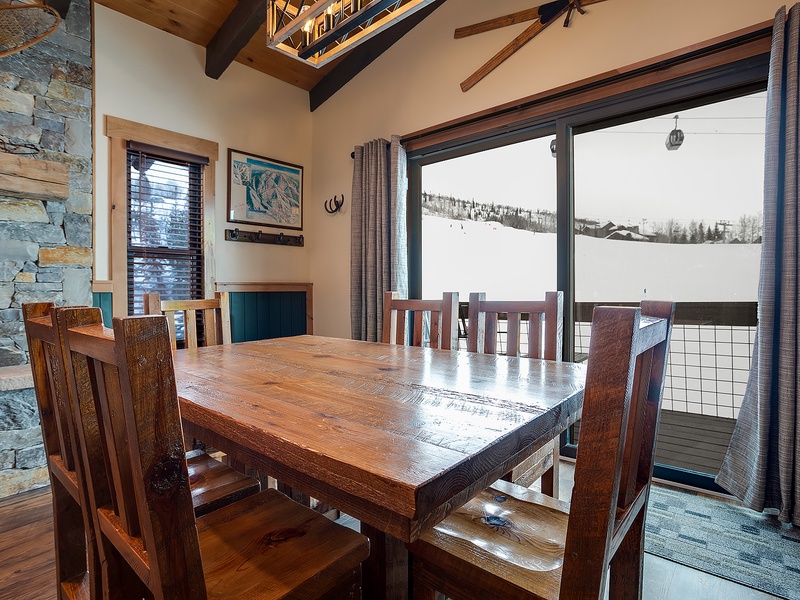 Gather around the handcrafted wooden table in your dining area, where floor-to-ceiling windows frame breathtaking ski slope views. The vaulted ceiling, stone fireplace, and mountain scenery create an unforgettable alpine retreat ambiance.