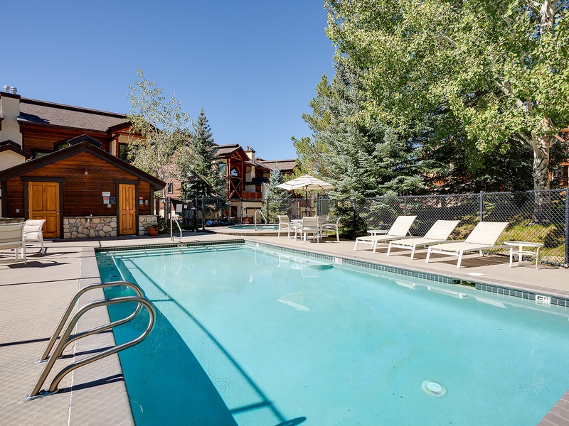 The property features a seasonal outdoor pool surrounded by mountain pines and deck chairs, with a wooden cabin and alpine-style buildings visible around the courtyard.