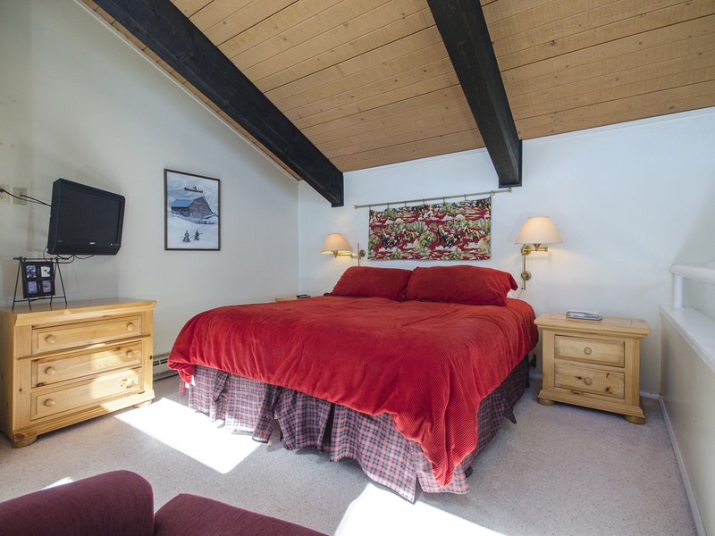 Drift off under charming exposed beams with warm bedside lamps casting a soft glow. Your cozy retreat features a plush red bedspread, mountain-inspired art, and entertainment at your fingertips.
