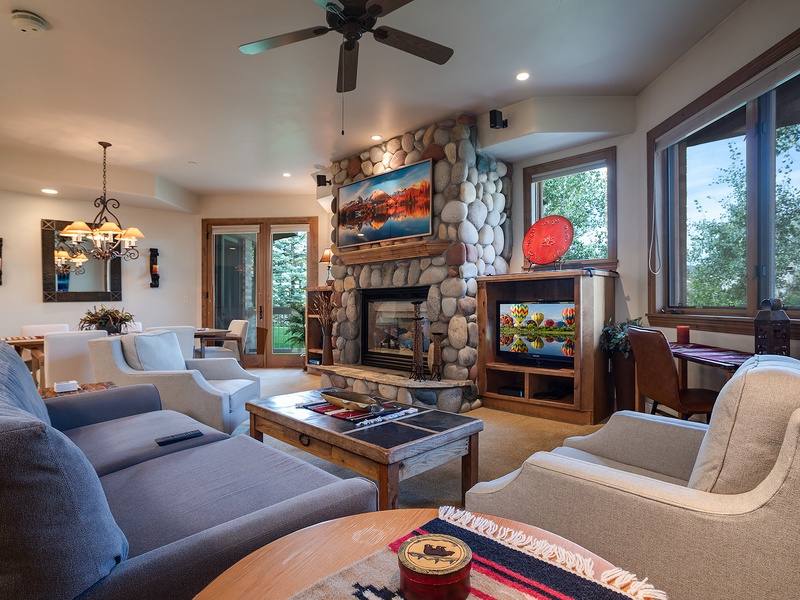 Relax in your spacious living room featuring a stunning stone fireplace, ceiling fan, and picture windows framing beautiful treetop views. Thoughtfully furnished with plush seating and warm lighting for effortless comfort.
