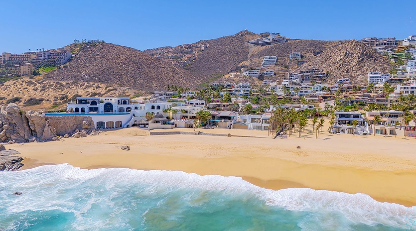 Beachfront hillside neighborhood with residential properties overlooking sandy shore and turquoise ocean waters, offering a scenic coastal location.