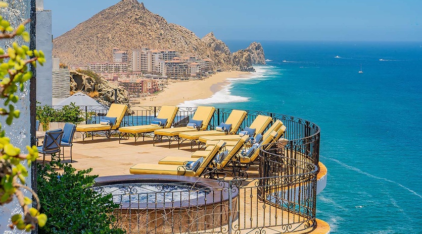 Elevated terrace featuring sun loungers and coastal panorama with dramatic rocky headland, pristine beach, and turquoise waters dotted with boats in the distance.