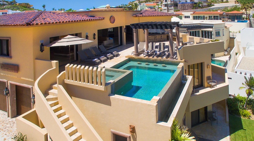 A luxurious hillside villa featuring a private rooftop pool with lounge chairs and shaded cabana, set against scenic neighborhood views and clear blue skies.