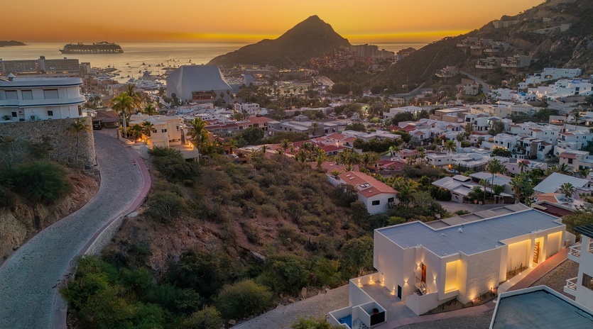 Stunning coastal hillside community features modern white villas overlooking azure waters during golden hour, with dramatic mountain backdrop and cruise ship views.