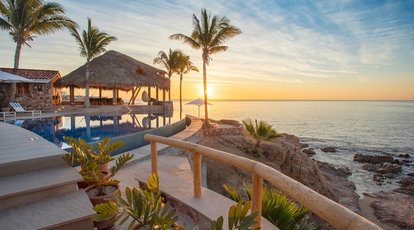 Beachfront property with infinity pool, thatched palapa, and palm trees overlooking the ocean at sunset, offering direct waterfront access and tropical coastal scenery.