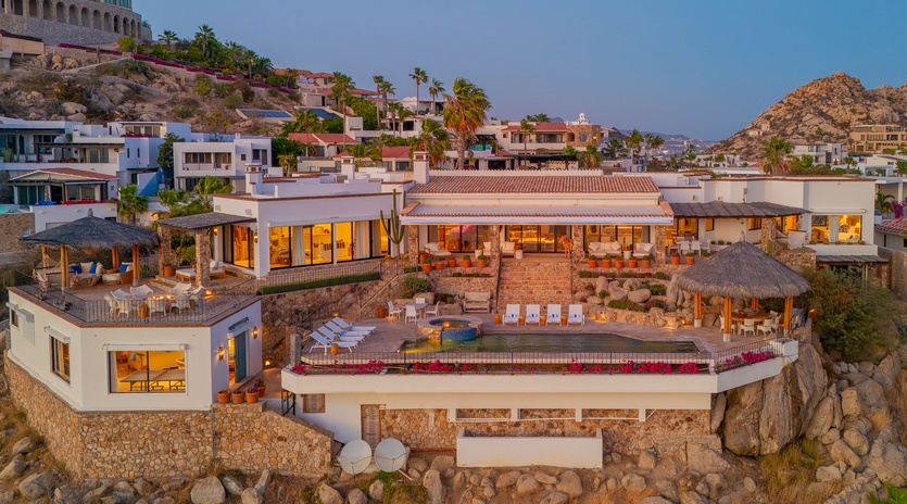 Luxury hillside villa perched on dramatic rocky coastline, featuring multiple levels with pools, terraces and thatched-roof palapas overlooking the sea.