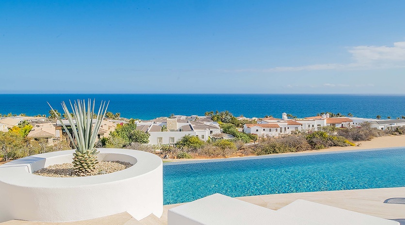 The infinity pool stretches toward the brilliant blue ocean, framed by sun-bleached architecture and desert plants—your perfect spot to soak up the Mediterranean sunshine.