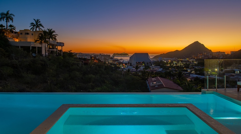 Stunning infinity pool overlooking tropical coastline at sunset, with dramatic mountain silhouettes and vibrant orange-purple sky creating the perfect evening backdrop.