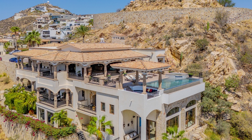 Stunning hillside villa showcasing Mediterranean architecture with rooftop pool amid dramatic desert landscape and neighboring coastal community.