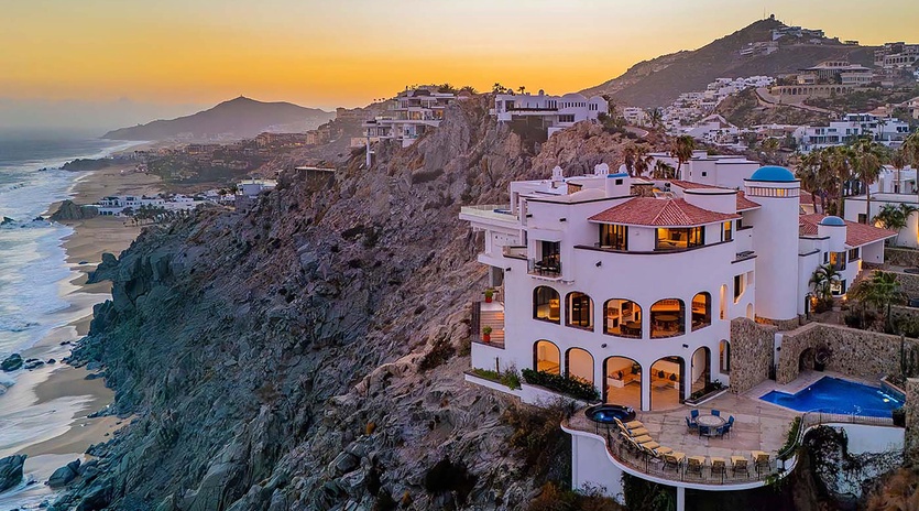 A stunning coastal estate perched on dramatic cliffs overlooking the ocean, captured during a breathtaking sunset. The property features hillside architecture, a private pool, and panoramic views of the beach and surrounding mountains.