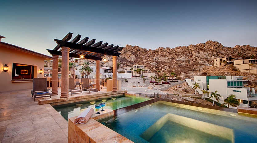 A dramatic hillside property with a multi-level pool and pergola-covered terrace, set against rugged desert mountains at dusk.