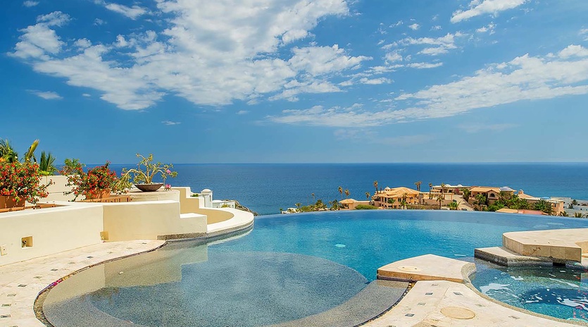 An expansive infinity pool overlooks the coastline, where sparkling blue water meets clear skies and neighboring oceanfront properties dot the landscape.