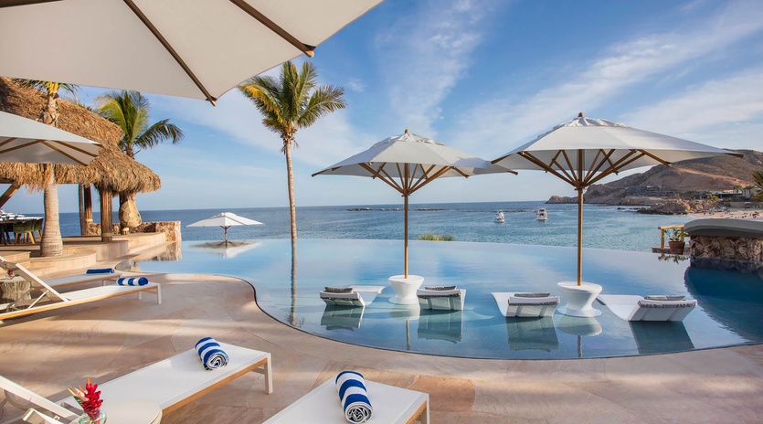 An infinity pool overlooking the ocean, featuring in-water lounge chairs under white umbrellas. Relax poolside with tropical palms swaying and coastal views stretching to the horizon.