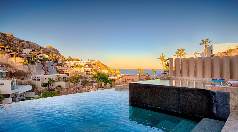 An infinity pool overlooking a hillside coastal community during golden hour. The property sits among terraced homes carved into the rocky landscape, with ocean views stretching to the horizon.