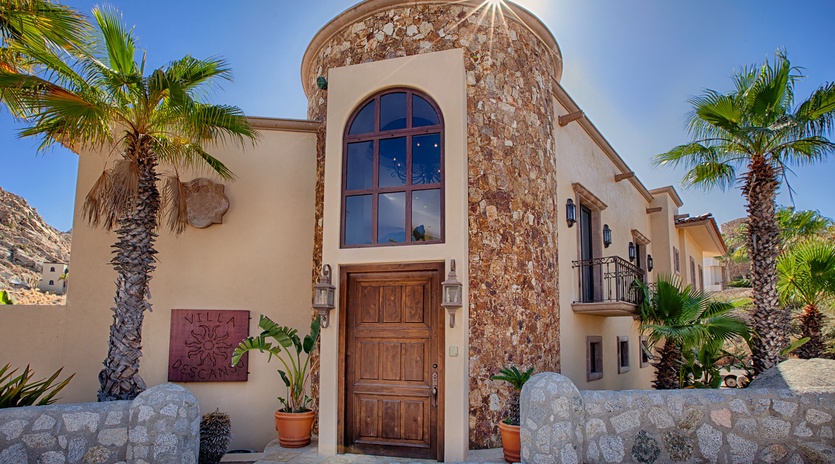 Mediterranean-style villa with distinctive stone turret entrance, arched wooden door, and palm-lined courtyard in a hillside setting.