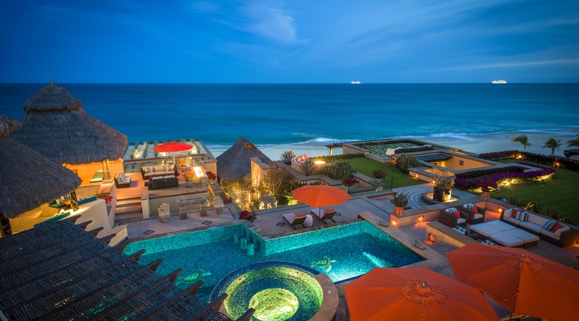 Spectacular beachfront villa complex featuring illuminated pool, hot tub, and luxury outdoor spaces with ocean views.