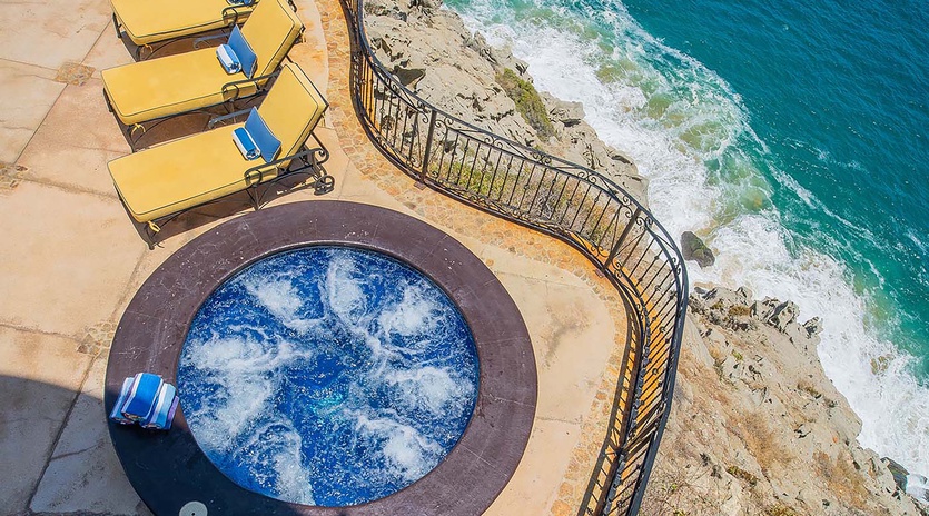 Aerial view of the property's dramatic oceanfront location, featuring a circular hot tub, yellow sun loungers on a curved terrace, and direct access to the rocky coastline and turquoise waters below.