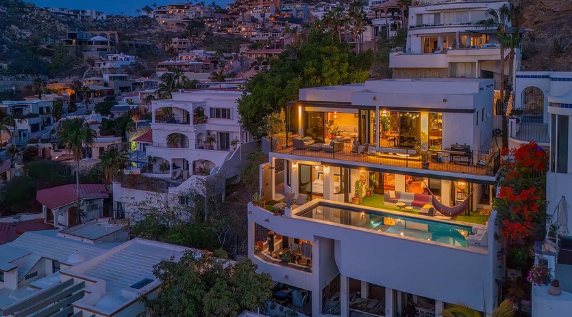 Hillside luxury villa with illuminated terraces and infinity pool at twilight, overlooking the neighboring coastal community and surrounding landscape.