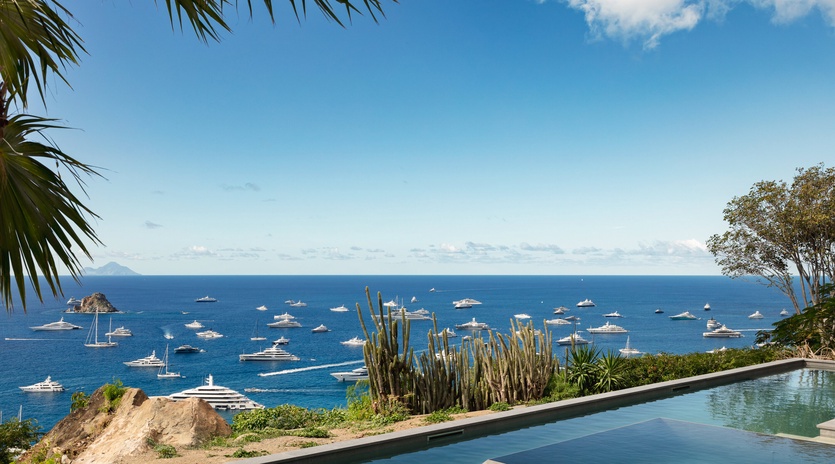 Infinity pool overlooking crystal-clear Mediterranean waters dotted with luxury yachts, framed by tropical gardens and distant island peaks.