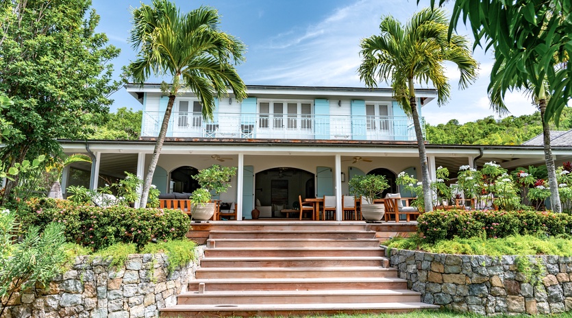 A charming Caribbean-style villa nestled among swaying palms and lush tropical gardens under bright blue skies.