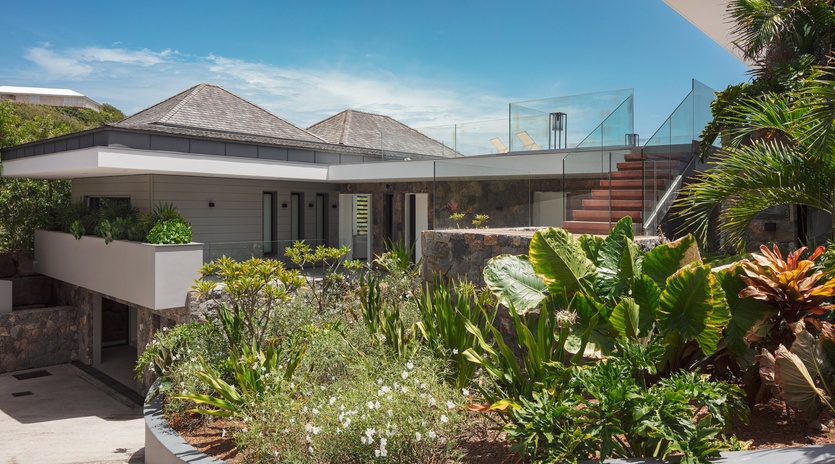 Modern tropical villa featuring contemporary architecture surrounded by lush landscaping and stunning glass-walled rooftop terrace under clear Caribbean skies.
