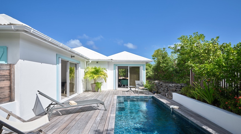 Modern white villa with private swimming pool surrounded by wooden decking and lush tropical landscaping under clear blue skies.