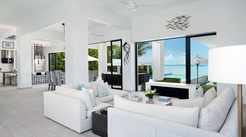 Sink into plush white sofas while turquoise waters and swaying palms frame your perfect tropical escape through floor-to-ceiling windows.