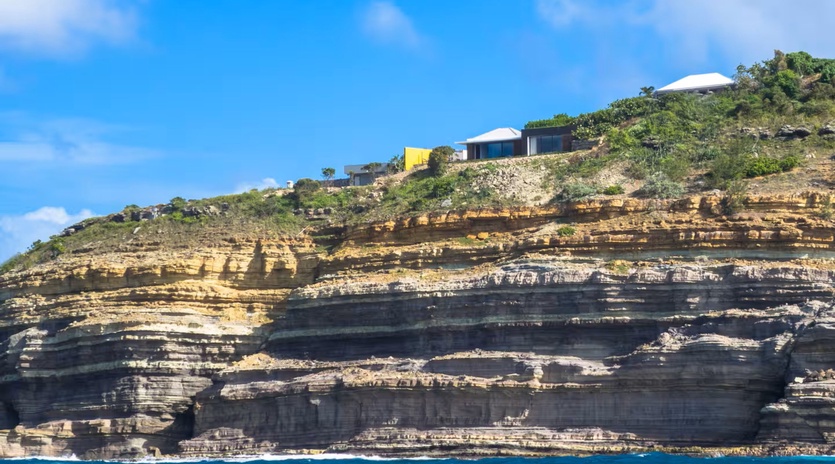 A striking hillside property nestled above dramatic coastal cliffs with layered sedimentary rock formations, lush greenery, and vibrant blue waters framing the scenic surrounding area.