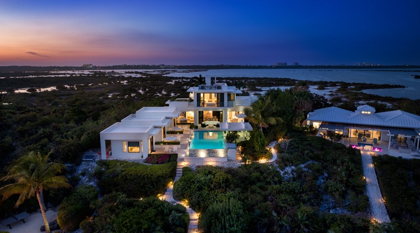 Aerial view of a modern waterfront estate at twilight, featuring a stunning pool, landscaped gardens, and expansive views over lagoons and natural surroundings.