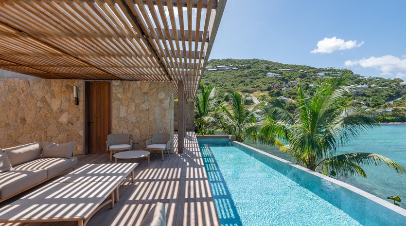 Your private infinity pool overlooks turquoise Caribbean waters, with tropical palms swaying and shaded lounging beneath elegant wooden pergola.