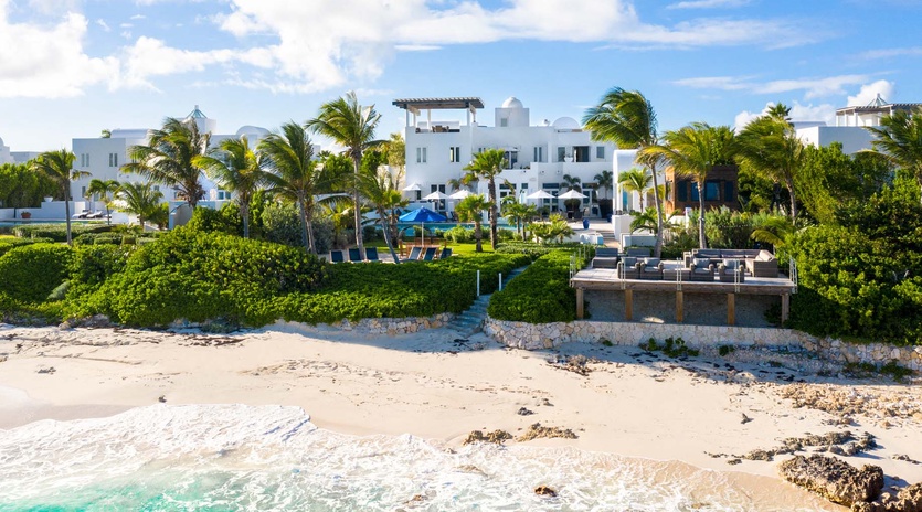 Stunning beachfront estate with white architecture, lush tropical gardens, and turquoise waters meeting sandy shores under bright blue skies.