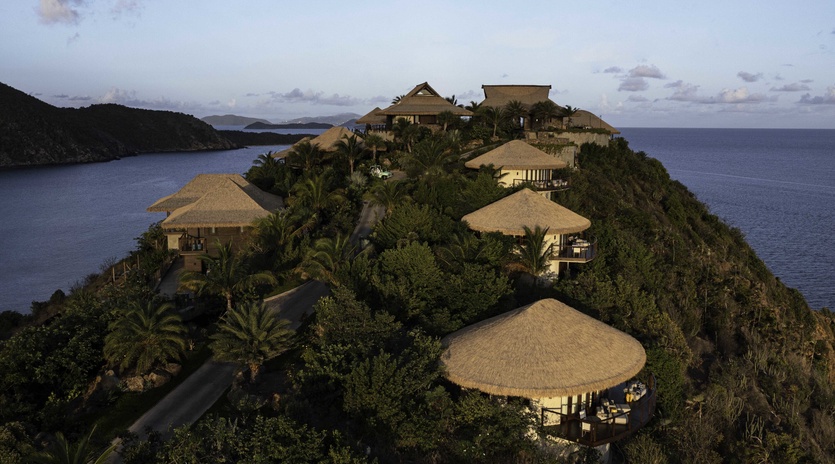 Tropical resort buildings perched dramatically on a lush hillside peninsula, surrounded by pristine blue waters and natural vegetation.