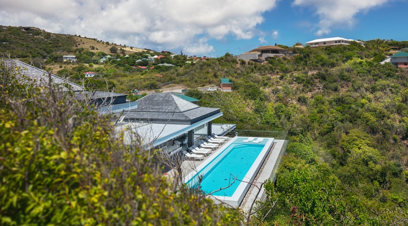 Modern hillside villa with pristine pool nestled among lush tropical vegetation and neighboring homes.