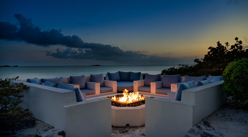 A stunning outdoor fire pit surrounded by built-in seating overlooks tranquil waters as twilight colors blend into night, providing an elegant beachside gathering spot.