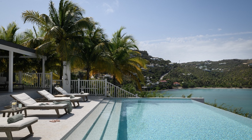 Stunning infinity pool overlooking turquoise bay waters, framed by swaying palms and hillside homes in this tropical paradise setting.
