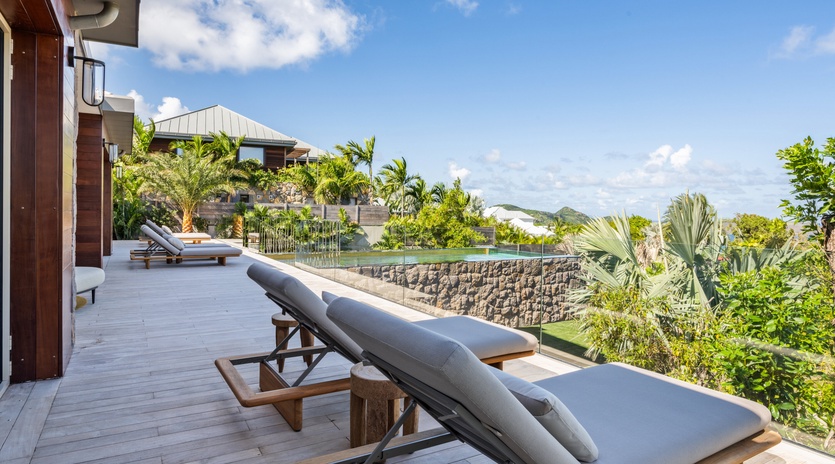 Spacious wooden deck with comfortable loungers overlooking tropical gardens and neighboring villas under bright blue skies.