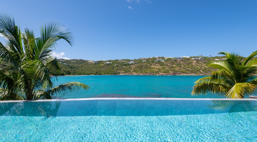 A stunning infinity pool creates the illusion of merging with crystal-clear turquoise waters beyond, framed by swaying palm trees.