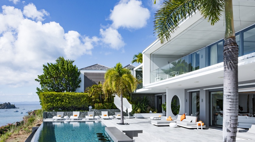 Modern luxury villa with infinity pool overlooking pristine waters, surrounded by tropical palm trees and contemporary architecture.