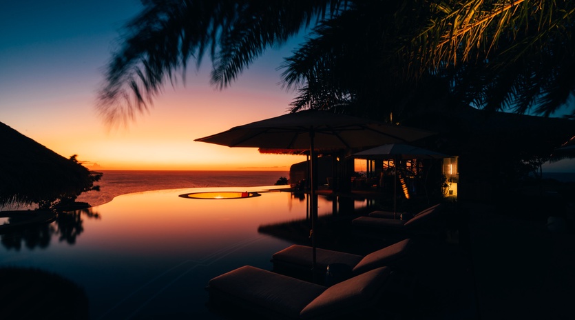 Tropical sunset paints the sky in brilliant oranges and blues over an infinity pool surrounded by swaying palms.