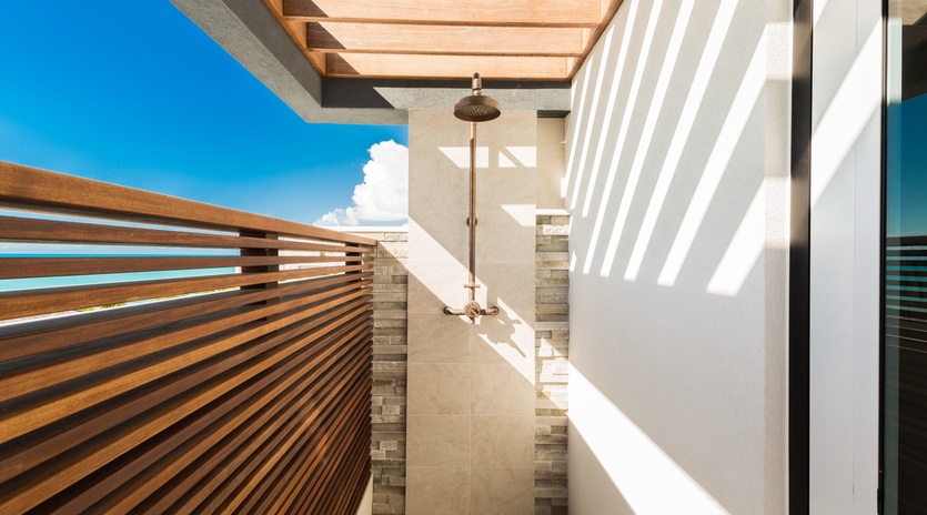 This stunning outdoor shower sits beneath a wooden-slatted pergola, open to clear blue skies. Clean stone walls and warm wood slats create a private, spa-like retreat.