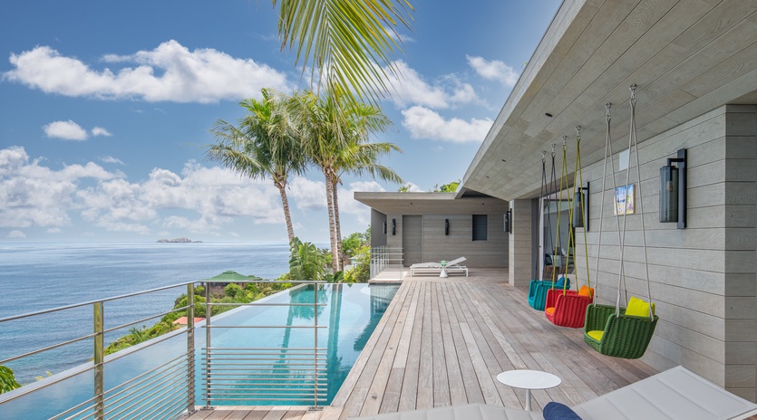 Modern oceanfront villa featuring infinity pool, tropical palms, and colorful swing chairs overlooking endless blue waters.