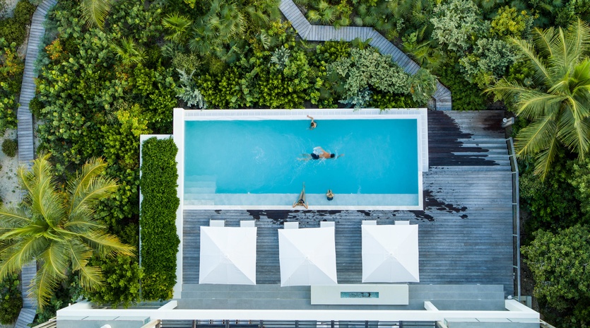 Stunning aerial view of a modern tropical villa featuring an infinity pool surrounded by lush rainforest and winding wooden walkways.