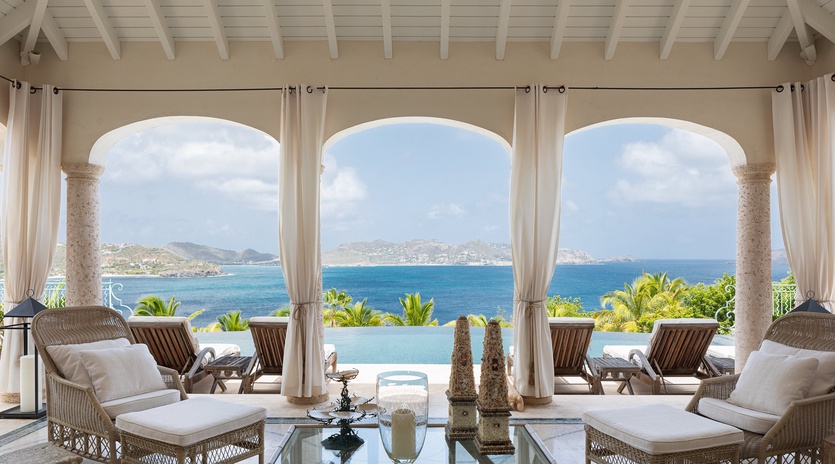Sink into plush wicker loungers and let your gaze drift across the sparkling turquoise waters and distant hillsides framed by elegant arches—your own private paradise.